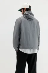 Campera rompevientos de nylon color gris oscuro, con forro de malla. Presenta cierre frontal con protección para el mentón, capucha ajustable con cordón, bolsillos laterales y un bolsillo interior, todos con cierre. Los puños y el bajo tienen elástico revestido.