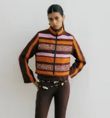 Chaqueta de cuello alto con cierre frontal, confeccionada en lino con diseño de rayas horizontales en tonos marrón oscuro, naranja y rosado. Presenta detalles de galones bordados con lentejuelas en el frente.