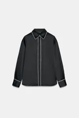 Camisa de corte amplio confeccionada en tejido satinado, con cuello solapa y manga larga. Presenta detalles de vivos en contraste y cierre frontal con botones ocultos.
