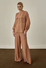 Camisa de gasa color capuchino, de manga larga con puños anchos abotonados, cuello mao y cierre frontal con botones dorados. Cuenta con bolsillos cargo en el pecho, detalles de charreteras en los hombros y un cinturón de tela ajustable con detalle de hebilla metálica dorada.
