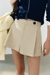 Short de tiro alto color marrón, con pliegues frontales y detalle de botones decorativos en la cintura. Cuenta con bolsillos laterales y cierre frontal con cremallera y gancho metálico.