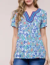 Blusa de manga corta con escote en V y pechera bordada. Presenta un estampado multicolor sobre base blanca, con pliegues frontales y aberturas laterales en el ruedo.
