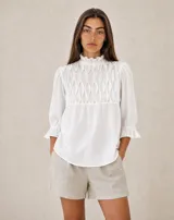 Blusa blanca de corte holgado, con cuello alto fruncido y detalle de nido de abeja en el frente. Cuenta con mangas 3/4 abullonadas con puños elásticos y volados.