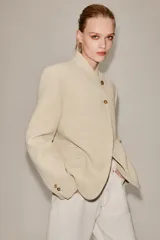 Blazer de corte estructurado en color beige, con cuello mao y cierre frontal cruzado mediante botones. Presenta mangas largas con detalle de botones en los puños y bolsillos de vivo en el frente.