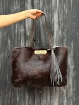 Cartera tipo bolso de mano y hombro de cuero con textura de pelo en color marrón oscuro, con herraje metálico dorado de cierre frontal y detalle de borla de flecos colgante.