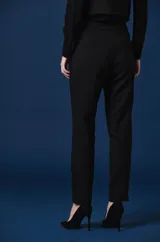Pantalon de vestir color verde oscuro, de corte recto y tiro alto. Presenta dos solapas decorativas en la cintura con apliques de broches metálicos con forma de insectos.