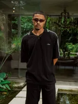 Chomba de tejido de punto cerrado color blanco, con cuello polo y mangas cortas. Presenta un bordado sutil en el frente y tres franjas negras en los hombros.