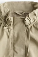 Campera corta de sarga de algodón, color beige, con cuello elevado ajustable por cordón, solapa rompevientos con botones a presión ocultos y cierre. Presenta bolsillos delanteros insertados, mangas ranglán con hombros caídos y bajo globo que genera una silueta abullonada.