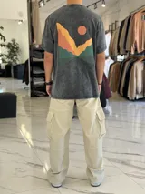 Remera oversize color negro con efecto gastado, manga corta y estampa artística en la espalda con diseño de montañas y sol en tonos amarillos, naranjas y verdes.