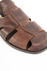 Sandalia tipo cangrejera de cuero marrón con diseño de tiras entrelazadas, puntera cerrada y cierre con hebilla metálica en el tobillo.