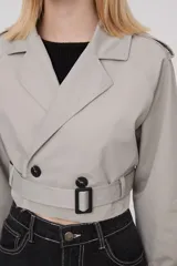 Trinchera corta color gris claro, con solapas, doble botonadura, cinturón y mangas largas con trabillas ajustables.