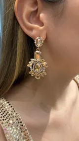 Caravanas colgantes de diseño elaborado con base dorada, compuestas por un cristal superior ovalado rodeado de strass y una pieza principal con forma de flor con pétalos de cristales en tonos ámbar y centro de gota, rodeado de pequeños cristales brillantes.