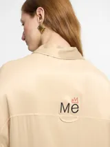 Camisa color beige de tejido satinado, corte oversize, manga larga, tapeta con botones invisibles y logo bordado en la espalda.