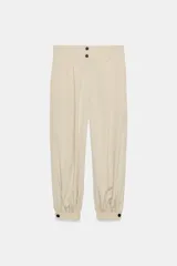 Pantalón de tiro alto en color beige, con diseño de pierna holgada y puños elásticos con cierre de botón en el bajo. Presenta cierre frontal con cremallera y doble botón en la cintura, además de bolsillos laterales.