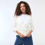 Sweater de punto color blanco con diseño cruzado tipo cárdigan, escote en V profundo y cierre lateral con un botón dorado. Presenta mangas largas con puños ajustados y un corte entallado a la cintura.