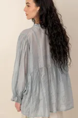 Blusa de seda gris con mangas largas abullonadas y puños con volados, escote en V con cuello fruncido y botones frontales.