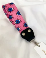 Llavero de muñeca con cinta de tela estampada con ojos azules sobre fondo rosado, conector de cuero sintético negro y gancho metálico plateado.