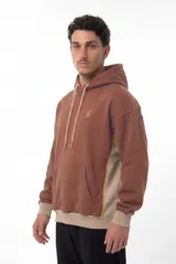 Hoodie oversize color marrón con paneles laterales en beige y detalles de costuras en contraste amarillo. Presenta capucha con cordones ajustables, bolsillo tipo canguro frontal y logo bordado en el pecho.