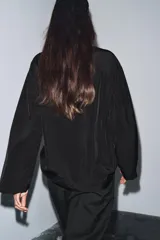 Blusa de cuello redondo y manga larga, confeccionada en tejido fluido con acabado satinado. Presenta un diseño de corte holgado, bajo recto y cierre en la espalda mediante una pequeña abertura con botón.