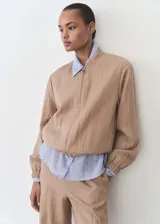 Chaqueta estilo bomber color beige, con cuello de tira, cierre frontal de cremallera, bajo y puños elásticos fruncidos y dos bolsillos laterales.