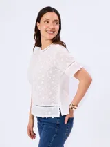 Blusa blanca de algodón con delicados bordados florales, cuello redondo y mangas cortas. Presenta detalles de calados geométricos en las costuras y aberturas laterales en el dobladillo.