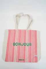 Bolso tote de tela con estampado de rayas verticales rosadas y blancas, laterales y asas en color beige. Presenta la palabra "BONJOUR" en verde en el centro y "INDIAN" en la parte inferior.