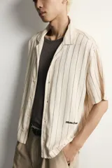 Camisa relaxed fit de manga corta y cuello bowling, confeccionada en tejido fluido en tono beige con diseño de rayas verticales oscuras. Presenta bordados a contraste y cierre frontal con botones.