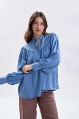 Blusa de jean de corte holgado en color azul claro, con cuello alto fruncido, lazo de ajuste en el escote y mangas largas abullonadas con puños abotonados.