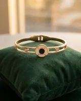 Esclava rígida de acero con baño de oro, diseño calado con detalle circular central en nácar y ónix, decorada con dos circonias. Incluye una cadena fina adicional integrada.