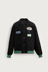 Cazadora varsity de pana de algodón negra, ligeramente acolchada, con cuello solapa, mangas largas y cierre frontal de botones a presión. Presenta parches bordados decorativos en el pecho y elástico rayado verde, blanco y negro en los puños y el ruedo.
