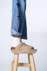 Zueco de gamuza sintética color beige, con detalle de tachas metálicas doradas y hebilla lateral ajustable. Presenta una base de corcho y suela de goma dentada.