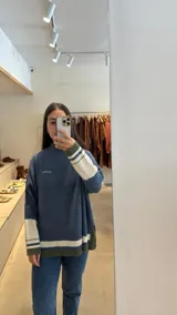 Buzo tejido de punto con cuello alto, diseño holgado y aberturas laterales. Presenta un cuerpo en color azul melange con detalles de franjas en color crema y verde en los puños y el ruedo. Incluye un pequeño bordado en el pecho.