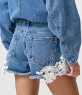 Short de jean tiro medio con encaje floral blanco aplastado en los laterales y dobladillo deshilachado. Cuenta con bolsillos delanteros y traseros, pasacintos y cierre frontal con botón y cremallera.
