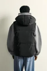 Chaleco regular fit acolchado para hombre, confeccionado en tejido técnico negro con acabado efecto engomado. Cuenta con cuello subido, capucha ajustable, bolsillos de vivo en la cadera, bolsillo interior y bajo ajustable con elástico en los laterales. El cierre frontal es con cremallera.