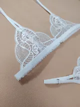 Conjunto de lencería de encaje blanco, compuesto por un corpiño triangular de tirantes regulables y una bombacha tipo colaless con tiras laterales ajustables.