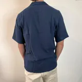 Camisa de hombre color verde oliva, de muselina con textura arrugada, manga corta, corte oversize, cuello cubano y cierre con botones. Presenta un pequeño logo bordado en el pecho.