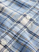 Camisa de manga larga confeccionada en tela de franela con estampado de cuadros en tonos azul claro, blanco y negro. Presenta cuello clásico, cierre frontal con botones y un bolsillo en el pecho.