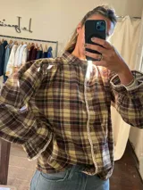 Camisa de manga larga con estampado a cuadros en tonos marrones y beige. Presenta cuello alto, cierre frontal con botones y detalles de puntilla blanca en la tapeta y en los puños.