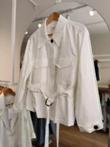 Campera blanca de algodón con cuello amplio, bolsillos delanteros con solapa, mangas largas y cinturón ajustable con hebilla redonda oscura. Cuenta con botones negros a contratono.