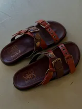 Sandalias de cuero color marrón, con diseño de tiras cruzadas, hebillas metálicas y tachas decorativas.