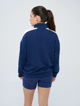 Buzo de felpa azul marino con medio cierre, cuello alto, bandas laterales en tono rosado y bolsillo frontal tipo canguro. Presenta el logo de Umbro y el escudo del Club Nacional de Football bordados.