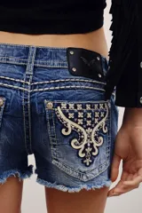 Short de denim azul de tiro bajo, con lavado desgastado, dobladillo desmechado y bolsillos traseros con bordados ornamentales y tachas metálicas.