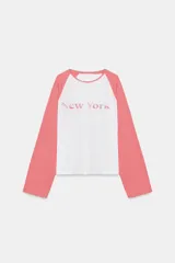 Camiseta de manga larga tipo ranglán con cuerpo blanco y mangas en color rosado. Presenta un estampado frontal con la inscripción 'New York' en tono rosado.