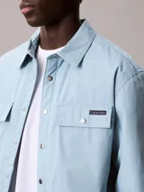 Sobrecamisa de algodón de corte regular, color celeste, con cierre frontal de botones a presión, bolsillos frontales con solapa y puños abotonados. Presenta un parche con el logo de la marca en el bolsillo superior izquierdo.