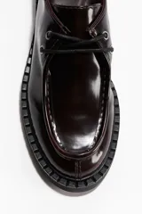 Zapato de vestir de cuero charol color marrón oscuro (casi negro), estilo mocasín con costura decorativa en la puntera. Posee cordones con ojales metálicos y suela gruesa de goma negra con tacón bajo y cuadrado.