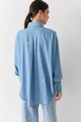 Camisa de jean azul claro con bordados pequeños en color blanco de figuras deportivas. Presenta cuello clásico, cierre frontal con botones de madera y mangas largas con puños abotonados.