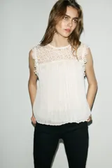 Top sin mangas de color blanco con cuello redondo. Presenta un panel superior de encaje tipo crochet y cuerpo plisado con forro interior. Cierre en la espalda mediante abertura con botón.