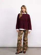 Sweater de punto oversize en color bordó con destellos de lurex. Presenta un diseño acanalado vertical, cuello redondo y mangas largas con terminaciones acampanadas y bordes ondulados.