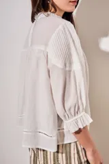 Blusa de algodón peinado color off-white, con cuello mao, cierre frontal de botones, mangas tres cuartos abullonadas con puños con volados, detalles de alforzas y apliques de encaje en el frente y los hombros.