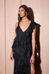 Vestido largo negro con escote en V y tirantes finos. Presenta volados verticales en cascada a lo largo del cuerpo y un tajo pronunciado en la parte delantera de la falda. La espalda es baja con tiras ajustables.